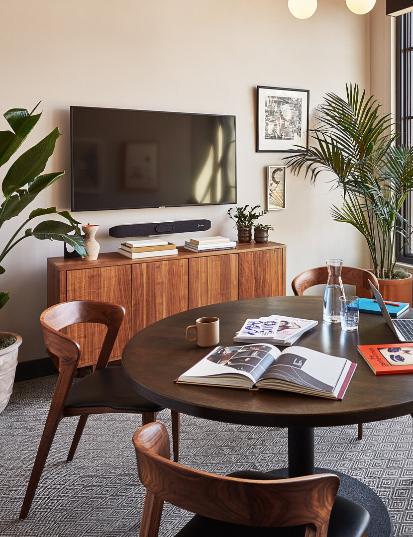 An office with a round wooden table with books and a laptop casually laid out on it. There is a TV on the wall and two large plants either side.
